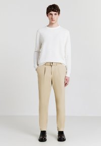 Maglione lavorato bianco abbinato a pantaloni beige plissettati. Stivali neri alla caviglia completano l'outfit su uno sfondo chiaro.