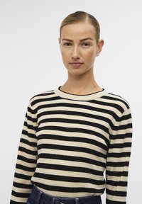 Pull en tricot noir et crème à rayures, avec un col rond et des manches longues, offrant une texture douce et une silhouette ajustée.