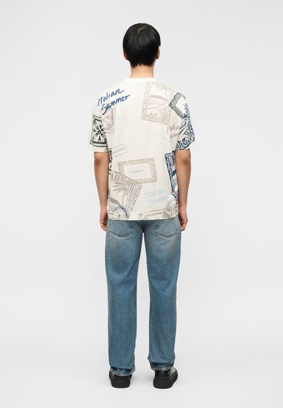 Άνδρας γυρισμένος προς τα πίσω, φοράει λευκό γραφικό T-shirt με το κείμενο «Italian Summer» και μπλε τζιν, μπροστά σε ένα απλό φωτεινό φόντο.