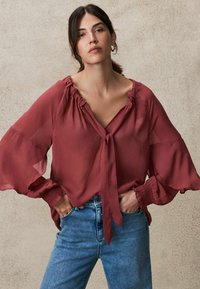 Bordeauxfarbene, leichte Bluse mit rüschigem Kragen und weiten, ausgestellten Ärmeln. Mit einem Front-Knotendetail und getragen mit blauen Jeans.