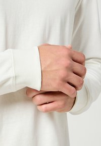 Biała koszula z długim rękawem z prążkowanym mankietem, z złożonymi dłońmi. Miękka, gładka tkanina z subtelną fakturą.