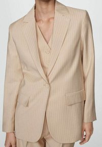 Veste de costume beige à fines rayures avec revers crantés, fermeture à un bouton et deux poches avant. Texture lisse, coupe ajustée.