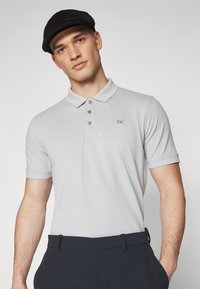 Svijetlosiva polo majica s klasičnom ovratnikom, tri gumba i CK logom. Nosi se uz tamne hlače, prikazujući ležeran, ali elegantan izgled.