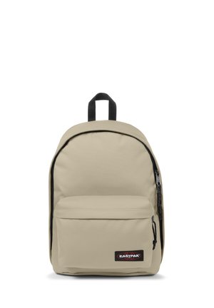 Mochila de tela beige con parte superior curvada, compartimento principal con cremallera y bolsillo frontal. Cuenta con un parche de logo negro y un bucle para colgar.
