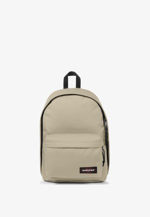 Mochila de tela beige con parte superior curvada, compartimento principal con cremallera y bolsillo frontal. Cuenta con un parche de logo negro y un bucle para colgar.