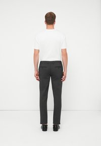 Egy férfi áll háttal, fehér pólóban, sötétszürke nadrágban, hátsó zsebekkel, és fekete cipőben egy sima fehér háttér előtt.