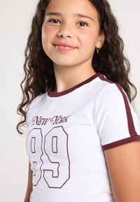 Ragazzina con capelli ricci che indossa una maglietta bianca con bordi bordeaux e la scritta "New York 99", sorridendo leggermente, su uno sfondo neutro.