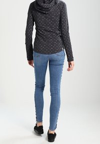 Kvinna i blå skinny jeans med svarta sidoknappar, mörk mönstrad långärmad topp och svarta avslappnade skor som står vänd bort.