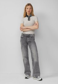 Lichtgrijze aansluitende geribde top, gecombineerd met grijze uitlopende jeans. Model draagt een zwart patroon sjaal en zilveren sneakers. Neutrale achtergrond.