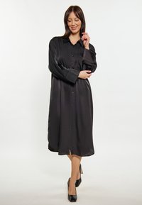 Schwarzes Satin-Hemdkleid mit Knopfverschluss auf der Vorderseite, langen Ärmeln, einem Kragen und seitlichen Schlitzen, kombiniert mit schwarzen High Heels.