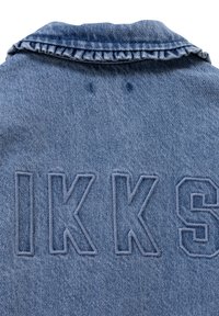Chaqueta de mezclilla en color azul claro, con cuello volado y "IKKS" bordado en letras negritas de un azul ligeramente más oscuro en la parte posterior.