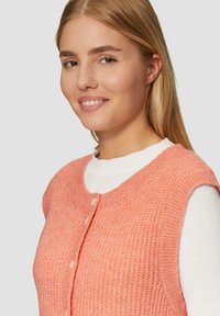 Pfirsichfarbene Strickweste mit strukturiertem Muster, runder Ausschnitt und Knopfleiste, über einem weißen Langarmshirt getragen.