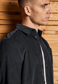 Camisa de pana negra con cuello y detalles de botones, y un bolsillo en el pecho. Textura suave con estrías verticales. El fondo presenta paneles de madera.