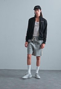 Young man wearing black cap and jacket, white tank top, gray shorts with "Palm Angels" text, white crew socks, and gray sneakers, standing indoors.