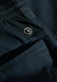 Pantalones cortos de tela azul marino con una cintura elástica, que presentan una pequeña etiqueta negra con un logotipo blanco en el lateral cerca de un bolsillo.