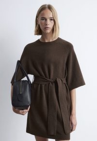 Braunes Strickkleid mit weiten Ärmeln und einem gebundenen Taillenbereich, kombiniert mit einer kleinen schwarzen Lederhandtasche mit goldfarbenem Reißverschluss und runder Form.