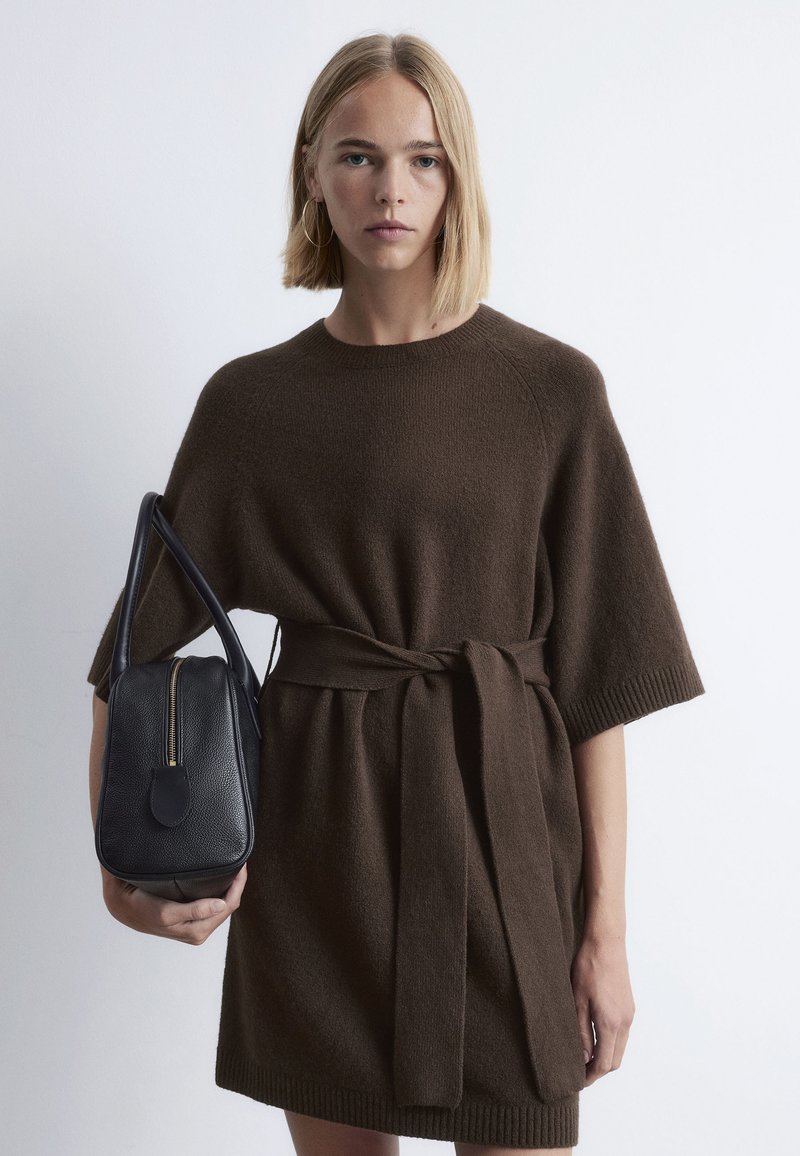Braunes Strickkleid mit weiten Ärmeln und einem gebundenen Taillenbereich, kombiniert mit einer kleinen schwarzen Lederhandtasche mit goldfarbenem Reißverschluss und runder Form.