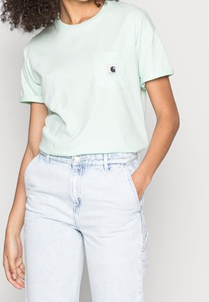 Persoon draagt een licht muntgroen shirt met korte mouwen en borstzakje en lichtblauwe, hooggetailleerde spijkerbroek, handen in de zakken.