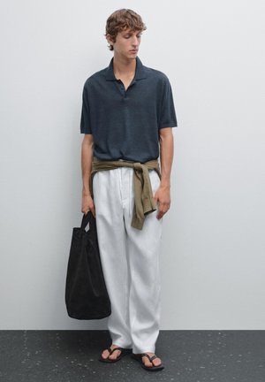Polo shirt blu scuro, pantaloni bianchi a vestibilità ampia, maglione olivastro annodato in vita, borsa a tracolla nera e infradito nere; texture lisce.