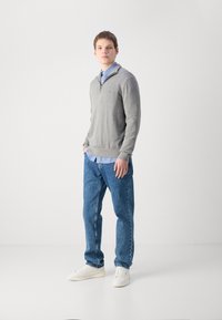 Pull gris avec col zippé superposé à une chemise bleu clair, assorti à un jean bleu et des baskets blanches, se tenant devant un fond uni.