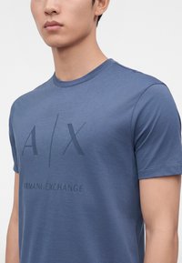 Άνδρας που φοράει μπλε κοντομάνικο T-shirt Armani Exchange με το λογότυπο A|X και το όνομα της μάρκας στο στήθος, εμφανίζεται από τους ώμους μέχρι τη μέση του κορμού.