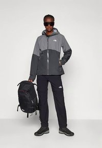 Chaqueta impermeable gris y negra con cremallera, combinada con pantalones negros y una mochila negra con detalles y logotipos rojos.