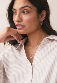 Silver heart pendant necklace on a delicate chain, worn with a striped shirt. Model's hand resting on her chin enhances visibility of the jewelry.