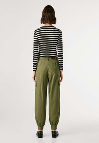 Vrouw met kort bruin haar, gekleed in een zwart-wit gestreepte lange mouwen top, olijfgroene cargobroek en zwarte schoenen, staande met haar rug naar ons gekeerd.