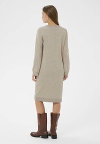Robe en tricot beige à manches longues, avec un col et un ourlet côtelés ; associée à des bottes en cuir marron hautes jusqu'au genou. Texture lisse, silhouette ajustée.