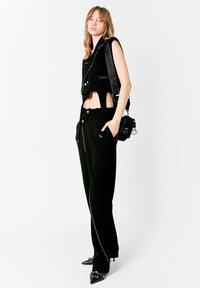 Top negro sin mangas con botones, pantalones negros de talle alto con cremalleras y tacones negros de punta puntiaguda. Pequeño bolso con correa.