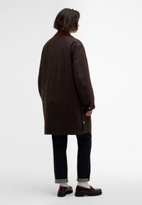 Barna, könnyű hosszú kabát, kontrasztos gallérral, patentgombokkal és enyhén túlméretezett fazonnal, sötét nadrággal és loaferekkel párosítva.