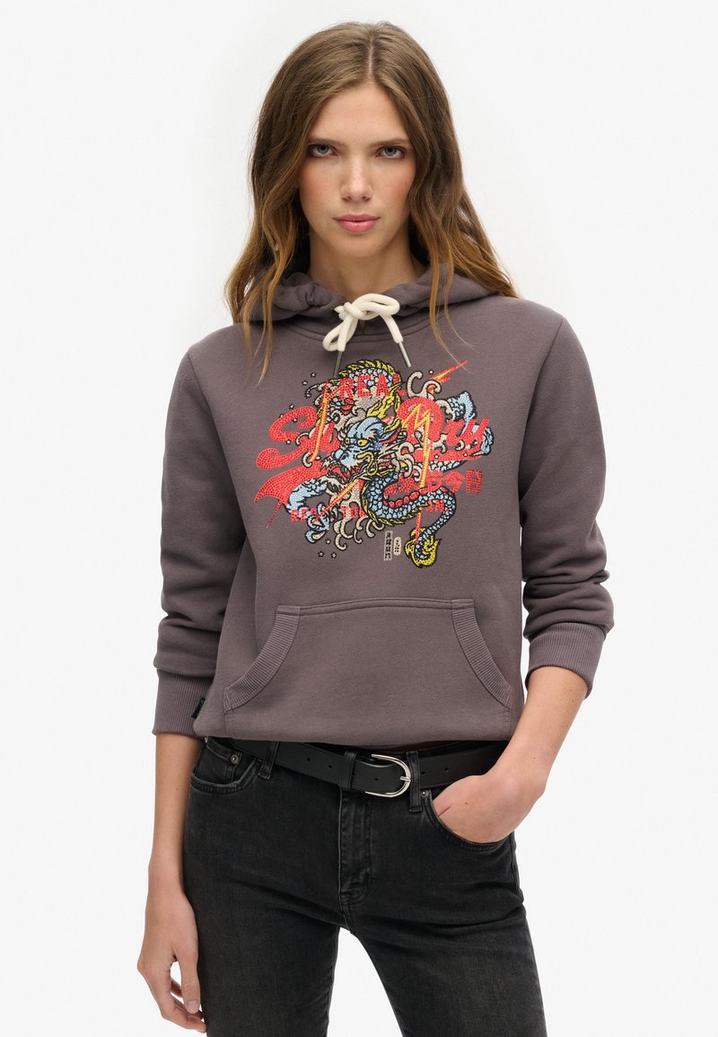 Młoda kobieta w szarej bluzie z kapturem z kolorowym graficznym wzorem smoka i sznurkiem, w połączeniu z czarnymi dżinsami i czarnym paskiem.