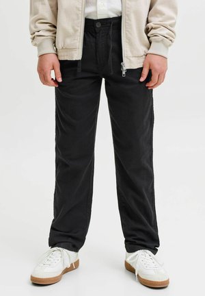 Persoon draagt zwarte loszittende broek, witte sneakers met rubberen zolen, beige jas met ritssluiting en wit overhemd, staand op een witte achtergrond.