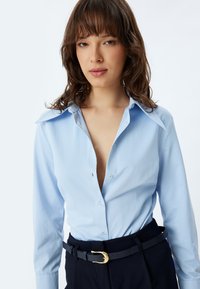 Camisa de manga larga de color azul claro con botón en la parte delantera y un cuello grande, combinada con pantalones oscuros y un cinturón negro con hebilla dorada.