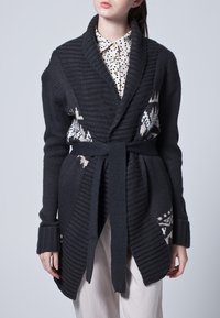 Ciemnoszary dziergany kardigan z kołnierzem szalowym, długimi rękawami i paskiem do wiązania. Na przodzie posiada białe geometryczne wzory.