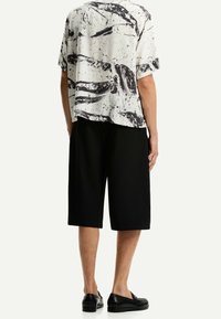 Lyhythihainen, väljähkö paita, jossa on valko-musta abstrakti kuvio, yhdistetty polvipitkiin mustiin shortseihin ja mustiin kenkiin.