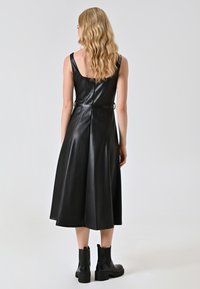 Schwarzes Leder-Midikleid mit runder Rückenausschnitt, verstellbarem Taillengürtel und glatter Textur. Kombiniert mit schwarzen Knöchelstiefeln.