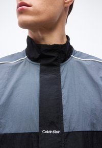 Κοντινό πλάνο ενός ατόμου που φοράει γκρι και μαύρο μπουφάν Calvin Klein με ψηλό γιακά, κοιτάζοντας ελαφρώς προς τα αριστερά.