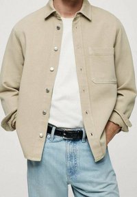 Chaqueta de camisa de algodón beige con botones a presión plateados, un bolsillo frontal y un corte relajado, combinada con una camiseta blanca y jeans de un azul claro.
