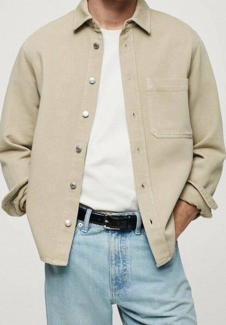 Chaqueta de camisa de algodón beige con botones a presión plateados, un bolsillo frontal y un corte relajado, combinada con una camiseta blanca y jeans de un azul claro.