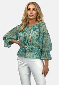 Turekzowy bluzka w kwiaty z falbaniastymi rękawami i elastycznym pasem, zdobiona wzorami paisley i kwiatowymi, zestawiona z białymi spodniami.