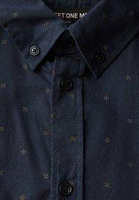 Tmavě modrá košile na knoflíky s jemnými malými béžovými geometrickými vzory a černými knoflíky podél légování a límce.