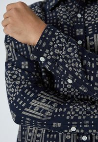 Tumedast sinistes mustriga särk, mis on valmistatud kangast, valgete geomeetriliste kujundite, nööbiga mansettide ja pehme tekstuuriga.