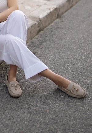 Beżowe zamszowe mokasyny z detalem w postaci kokardy, miękka faktura, zakładany fason i mały złoty akcent z przodu, w połączeniu z białymi szerokimi spodniami.