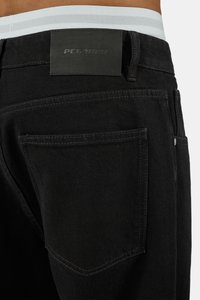 Rückansicht einer Person, die schwarze Jeans trägt, mit sichtbarer Gesäßtasche und schwarzem Pegador-Markenlabel am Bund.