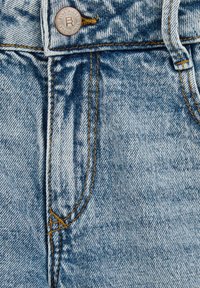 Primer plano de unos vaqueros de mezclilla azul desgastados, mostrando el bolsillo delantero, el lazo del cinturón, la costura anaranjada y el botón de metal etiquetado como "Blue Ridge."