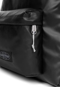 Sac à dos en nylon noir avec une poche zippée à l'avant, surface texturée et tirette argentée. Dispose d'un patch logo noir sur le côté.