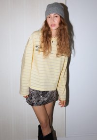 Sárga csíkos hosszú ujjú ing fekete felirattal, ezüst flitteres miniszoknyával párosítva. Szürke kötött sapka és fekete térdig érő csizma.