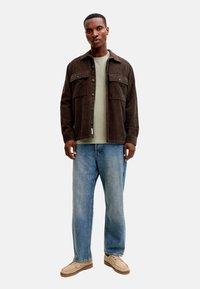 Άνδρας που φοράει ένα καφέ μπουφάν κοτλέ πάνω από ένα ανοιχτό πράσινο μπλουζάκι, μπλε τζιν και μπεζ καθημερινά παπούτσια, στέκεται με ουδέτερη έκφραση.