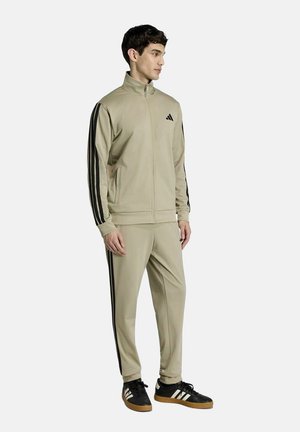 Uomo che indossa una tuta beige Adidas con strisce nere e scarpe da ginnastica Adidas nere, in piedi e guarda verso destra su sfondo semplice.